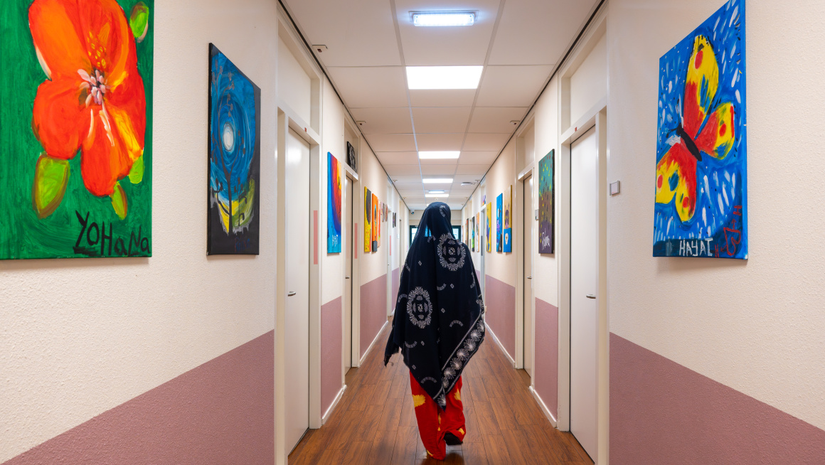 Jonge vrouwelijke asielzoeker in jurk loopt door de gang in het azc, we zien haar op de rug, er hangen kleurrijke schilderijen aan de muur