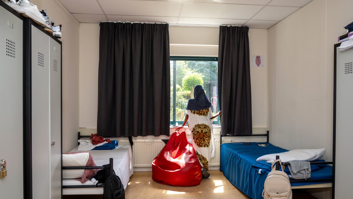 Jonge vrouwelijke asielzoeker in jurk kijkt uit het raam van haar slaapkamer in het azc Nunspeet. Er staan 2 opgemaakte bedden, 2 metalen kasten en een rode zitzak. 
