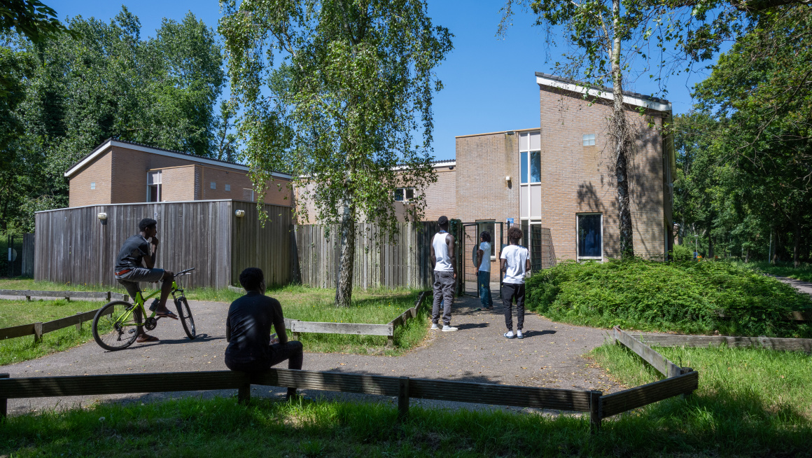 Minderjarige jongens staan en zitten voor de ingang van hun opvanglocatie in een groene omgeving.