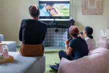 Drie amv meisjes zitten in de woonkamer. Ze kijken naar de tv en zingen mee met karaoke microfoons. Twee meisjes rechts zitten op een roze zitzak, het meisje links zit op een hoek van de grijze bank. De tv hangt boven de zwarte schouw met blauwe tegeltjes.